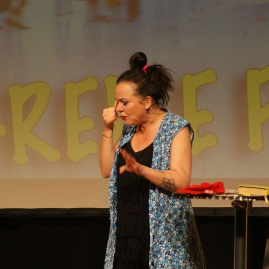Eine Frau auf der Bühne, die mit der Hand vor dem Mund performt, trägt ein schwarzes Kleid und eine Blumenweste. Hinter ihr ist ein Hintergrund mit dem Wort Erleichterung.