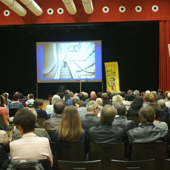 Eine Menschenmenge sitzt in Stühlen in einem Theater und schaut sich einen Film auf einer Leinwand an. Rechts ist ein Banner sichtbar.