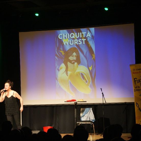 Eine Frau auf der Bühne singt in ein Mikrofon mit einer Leinwand im Hintergrund, die eine Bananenkarikatur zeigt. Die Leinwand zeigt 'Chiquita Wurst' und das Publikum ist im Hintergrund.
