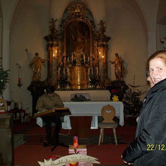 Eine Person spielt ein Musikinstrument in einer Kirche mit Weihnachtsdekoration. Eine Frau in einer schwarzen Jacke lächelt. Der Altar hat Kerzen, ein Gemälde und Statuen. Das Datum 22/12 ist auf dem Foto.