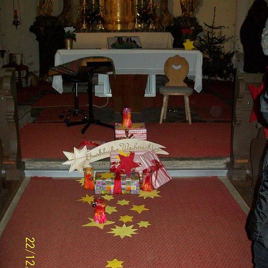 Eine Weihnachtsszene mit Geschenken und Sternen auf einem roten Teppich. Ein Holzstuhl und ein Schreibtisch stehen neben einem geschmückten Altar.