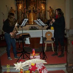 Zwei Frauen spielen Instrumente in einer Kirche, mit einem Weihnachtsbanner auf dem Boden. Ein Saxophon ist links und eine Klarinette rechts. Kerzen und Statuen sind hinter ihnen.