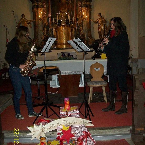 Zwei Frauen spielen Instrumente in einer Kirche, mit einem Weihnachtsbanner auf dem Boden. Ein Saxophon ist links und eine Klarinette rechts. Kerzen und Statuen sind hinter ihnen.