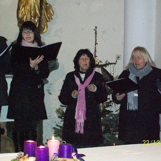 Vier Frauen in schwarzen Kleidern stehen neben einem geschmückten Weihnachtsbaum und singen aus einem Notenbuch. Dahinter ist eine goldene Statue an der Wand befestigt.