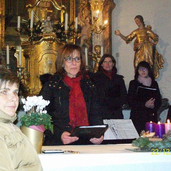Fünf Frauen stehen vor einem Kirchenaltar mit einer Statue, halten Bücher und singen. Eine Frau sitzt mit einem Blumentopf. Kerzen sind auf einem Schreibtisch angezündet.