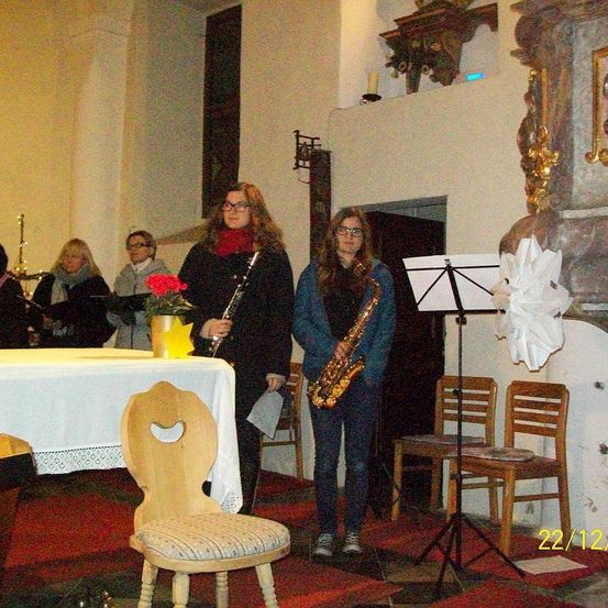 Eine Gruppe von Musikern steht in einem Raum mit Tisch und Stühlen. Zwei Frauen halten Instrumente, eine eine Saxophon und die andere eine Flöte. Eine Kerze ist an der Wand montiert.