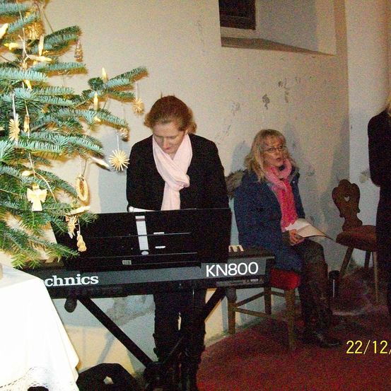 Zwei Frauen sind in einem Raum. Eine spielt Klavier, die andere sitzt auf einem Stuhl und liest. Ein Weihnachtsbaum steht hinter dem Klavier. Das Datum 22/12 ist auf dem Bild vermerkt.