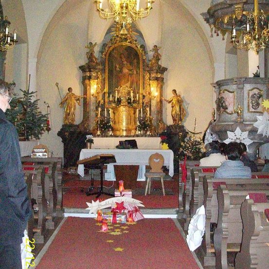 Im Inneren einer Kirche ist ein Altar mit Statuen und Kerzen geschmückt. Ein Mann steht am Eingang. Die Kirchenbänke sind mit Menschen gefüllt und der Gang ist mit festlichen Dekorationen geschmückt.
