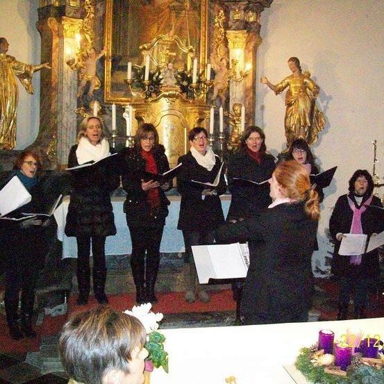 Ein Chor singt in einer Kirche vor einem geschmückten Altar. Die Chormitglieder tragen Winterkleidung. Ein Kerzenhalter mit Kerzen steht auf dem Altar.
