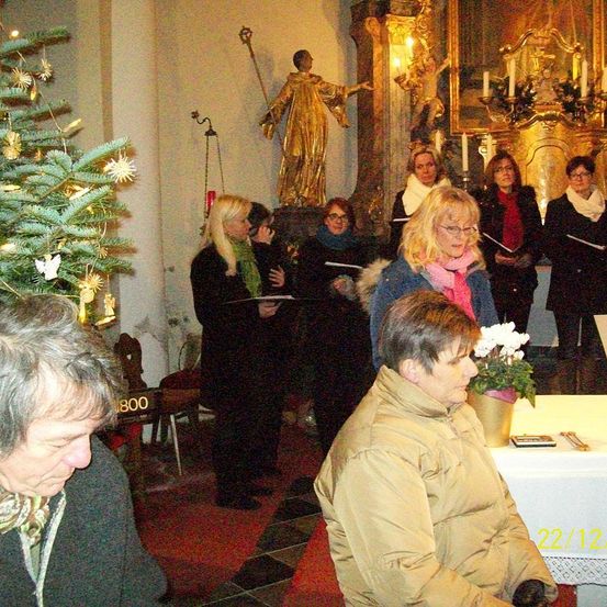 In einer Kirche singen eine Gruppe von Frauen und halten Bücher. Ein Weihnachtsbaum ist geschmückt. Eine goldene Statue eines Heiligen befindet sich an der Wand.