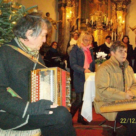 Zwei Musiker spielen Instrumente in einer Kirche, mit einem Weihnachtsbaum und einer goldenen Statue hinter ihnen. Eine Gruppe von Menschen steht im Hintergrund.