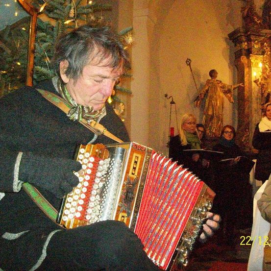 Ein Mann spielt Akkordeon bei einer Weihnachtsaufführung in einer Kirche. Hinter ihm singen Menschen, während eine Statue einer Heiligen im Hintergrund steht.