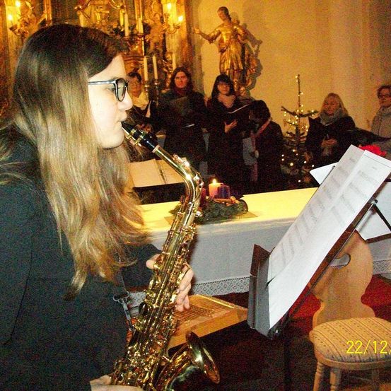 Eine Frau mit Brille spielt in einer Kirche Saxophon, mit einem Chor im Hintergrund. Es gibt einen geschmückten Tisch mit Kerzen und einer Statue hinter ihr.