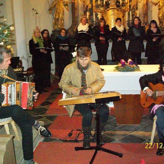 Eine Gruppe von Musikern spielt Instrumente in einer Kirche, während im Hintergrund ein Chor singt. Die Musiker umfassen eine Person, die Akkordeon spielt, eine Person, die Gitarre spielt, und eine Person, die Zither spielt.