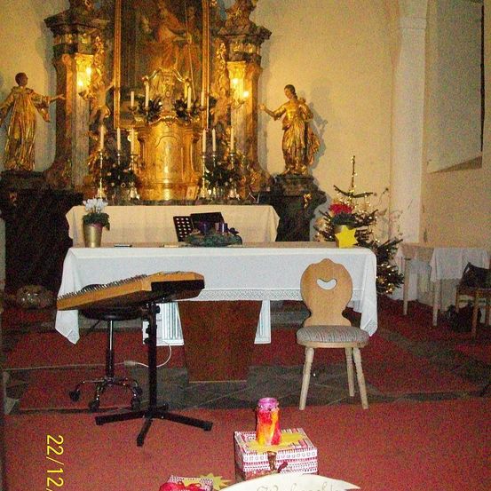 Ein goldener Altar in einer Kirche mit Kerzen, einer Statue und einem Weihnachtsbaum. Ein Holzstuhl und ein Notenständer stehen vor dem Altar. Ein Geschenkkarton liegt auf dem Boden.