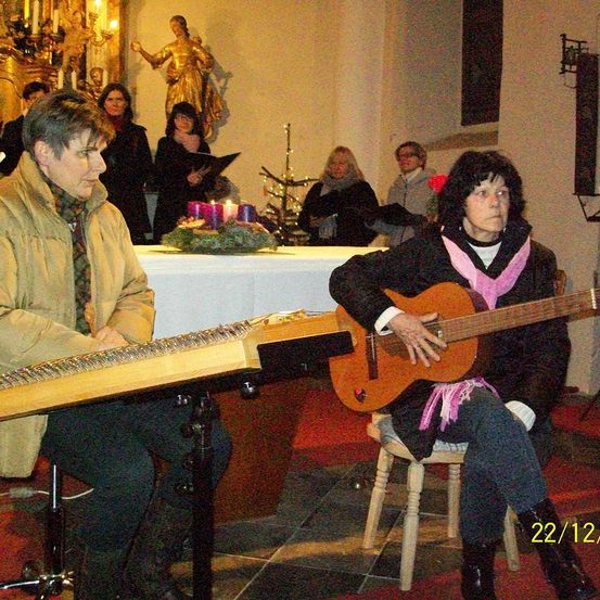 Zwei Frauen spielen Gitarren in einer Kirche, eine auf einem Hocker und die andere auf einem Ständer. Dahinter steht ein Chor hinter einem Tisch mit Kerzen. Eine Statue einer Frau ist an der Wand angebracht.