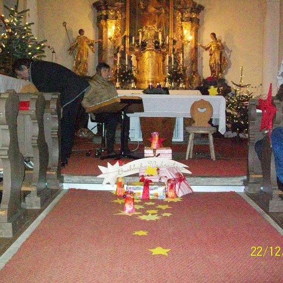 Ein Foto vom 22. Dezember zeigt eine geschmückte Kirche, in der zwei Personen an einem Tisch sitzen. Geschenke und Sterne liegen auf dem Boden, und im Hintergrund befinden sich zwei Statuen.