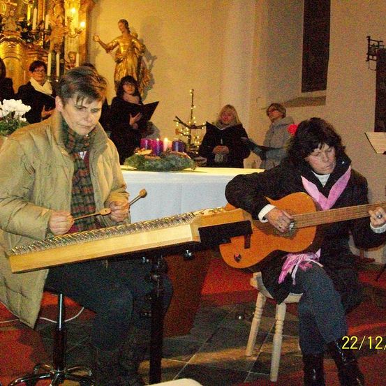 Zwei Frauen spielen Musikinstrumente in einer Kirche. Eine spielt Hackbrett, die andere Gitarre. Dahinter singt eine Gruppe von Menschen. Die Kirche ist für einen besonderen Anlass geschmückt.