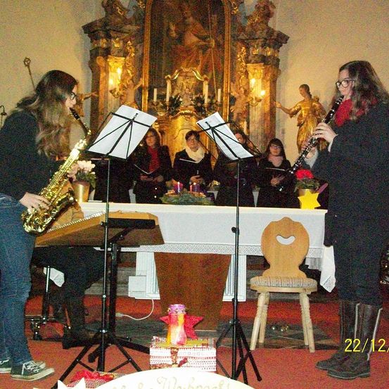 Zwei Frauen spielen Musikinstrumente in einer Kirche. Eine Frau spielt Saxophon, die andere Klarinette. Dahinter singt ein Chor mit Kerzen auf dem Altar. Die Szene zeigt einen Holzstuhl, einen Tisch und ein Gemälde an der Wand.