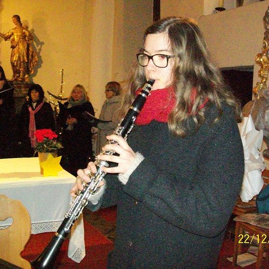 Eine Frau spielt Klarinette in einer Kirche, während andere singen. Ein Blumenvase steht auf einem Tisch vor ihr.