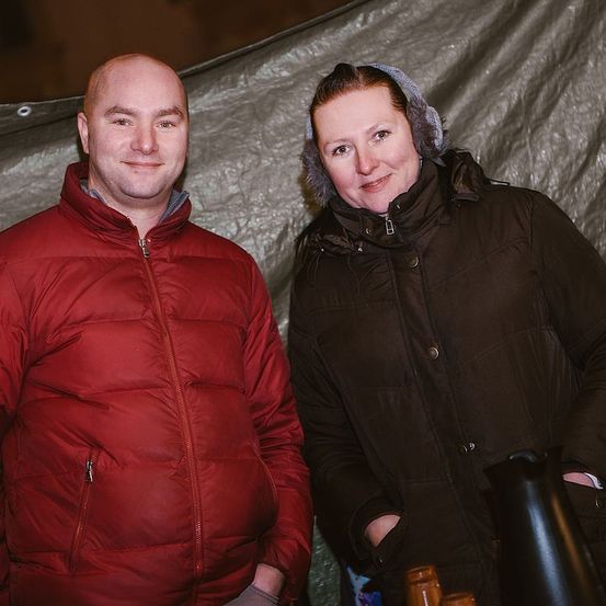 Ein Mann und eine Frau stehen Seite an Seite in Winterkleidung und lächeln. Der Mann trägt eine rote wattierte Jacke, und die Frau trägt einen braunen Mantel mit Kapuze. Hinter ihnen steht ein weißes Zelt.