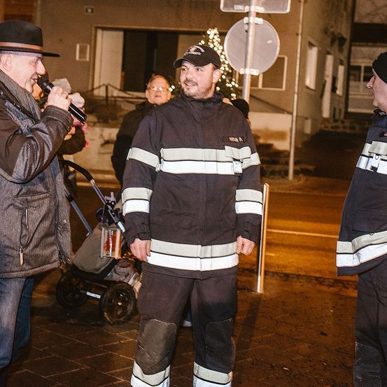 Ein Mann mit einem Mikrofon spricht, während ein Feuerwehrmann mit den Händen in den Taschen steht. Ein Kinderwagen mit einem Kind steht hinter ihnen. Ein Straßenschild ist im Hintergrund.