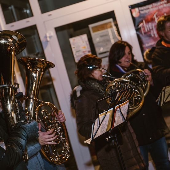 Eine Gruppe von Musikern, in Winterkleidung gekleidet, spielt französische Hörner in einem Raum mit Glastüren und einem Plakat an der Wand.