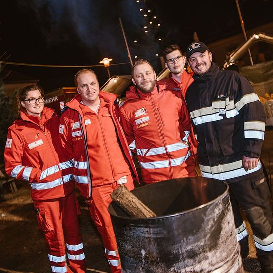 Fünf Personen in roten Uniformen, vermutlich Rettungskräfte, stehen neben einem Fass mit einem Baumstamm. Sie posieren für ein Foto bei Nacht, mit Gebäuden und Lichtern im Hintergrund.