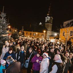 Eine große Menschenmenge versammelt sich bei Nacht im Freien. Viele tragen Winterkleidung und Mützen. Einige halten Becher und Flaschen. Gebäude und Bäume sind im Hintergrund.