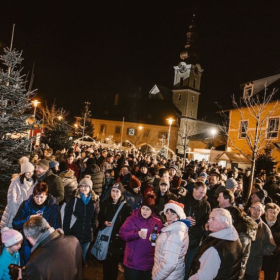 Eine große Menschenmenge versammelt sich bei Nacht im Freien. Viele tragen Winterkleidung und Mützen. Einige halten Becher und Flaschen. Gebäude und Bäume sind im Hintergrund.