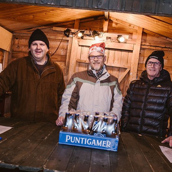 Drei Männer stehen lächelnd in einer Hütte. Einer trägt einen Weihnachtsmannhut, ein anderer hält eine Kiste Puntigamer Bier. Sie sind von Holzwänden umgeben, mit Deckenleuchten und einer Holztür.