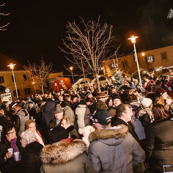 Eine Menschenmenge, eingehüllt in Winterkleidung, versammelt sich bei Nacht im Freien. Straßenlaternen und ein Gebäude im Hintergrund.