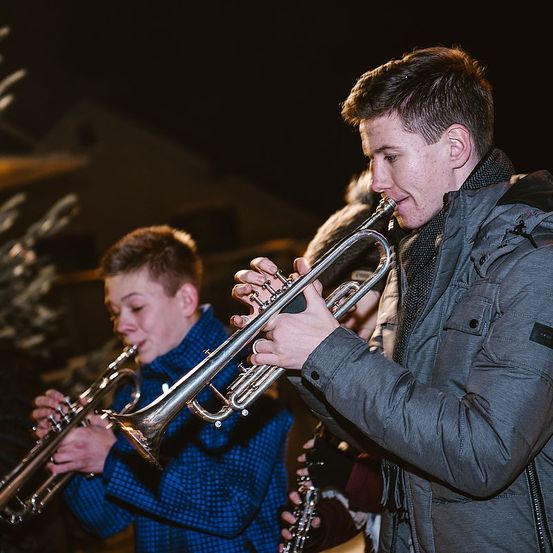 Zwei junge Männer spielen nachts Trompeten, wobei einer etwas voraus ist. Sie tragen beide Jacken, und der Mann vorn konzentriert sich auf das Spielen seines Instruments.