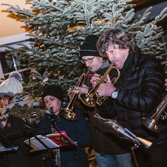 Eine Gruppe von Musikern spielt vor einem geschmückten Weihnachtsbaum. Sie spielen Trompeten und eine Klarinette, und einer von ihnen trägt eine Uhr.