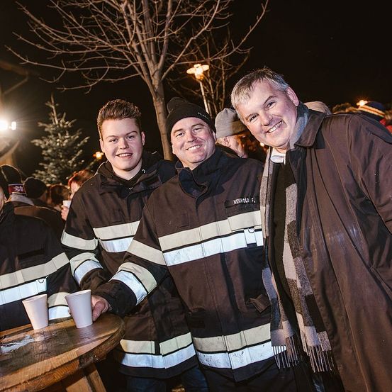 Drei Feuerwehrleute in Warnwesten stehen bei Nacht mit Tassen an einem Tisch. Der auf der linken Seite hält zwei Tassen. Sie lächeln. Bäume und eine Laterne sind hinter ihnen.