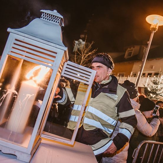 Ein Mann in einer Warnweste zündet eine Kerze in einer Laterne bei Nacht an. Er ist von Menschen umgeben, von denen einer ein Foto macht.
