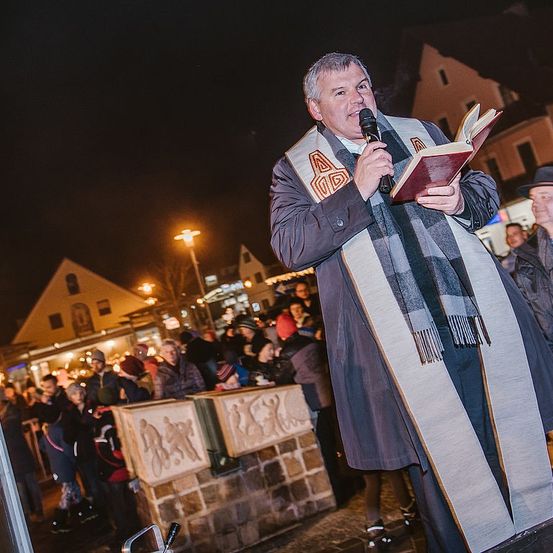 Ein Priester in einer Robe steht an einem Mikrofon und liest aus einem Buch. Hinter ihm versammelt sich eine Menschenmenge auf einem Platz. Im Hintergrund sind Gebäude und Straßenlaternen zu sehen.