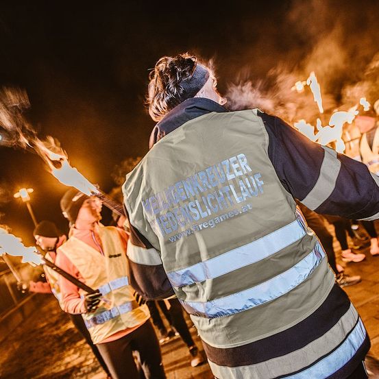 Eine Person in einer reflektierenden Sicherheitsjacke mit dem Text 'Verkehrskreuzer' steht bei Nacht mit anderen, die Fackeln halten. Sie nehmen wahrscheinlich an einer Fackelveranstaltung teil.