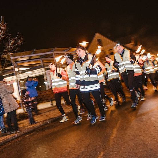 Eine Gruppe von Läufern mit Reflektorwesten und Helmen joggt bei Nacht auf einer Straße und hält Fackeln, mit Zuschauern an der Seite.