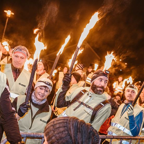 Teilnehmer einer nächtlichen Veranstaltung halten brennende Fackeln. Die Menge wird von den hellen Flammen beleuchtet. Der Mann vorne hat einen Bart und trägt eine reflektierende Weste.