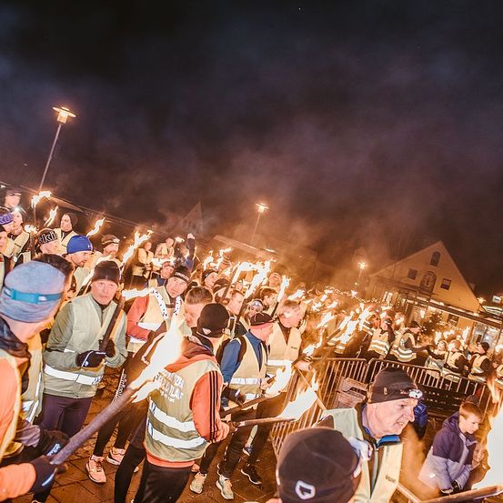 Eine Gruppe von Menschen, die in reflektierende Kleidung gekleidet sind, versammelt sich in der Nacht und hält brennende Fackeln. Sie scheinen sich auf eine Fackelprozession vorzubereiten. Rauch füllt die Luft.
