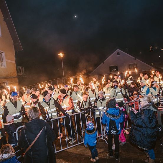 Eine Menschenmenge versammelt sich in der Dunkelheit und hält brennende Fackeln. Sie stehen hinter einem Metallzaun, einige tragen Warnwesten. Die Gebäude und Straßenlaternen sind im Hintergrund zu sehen.
