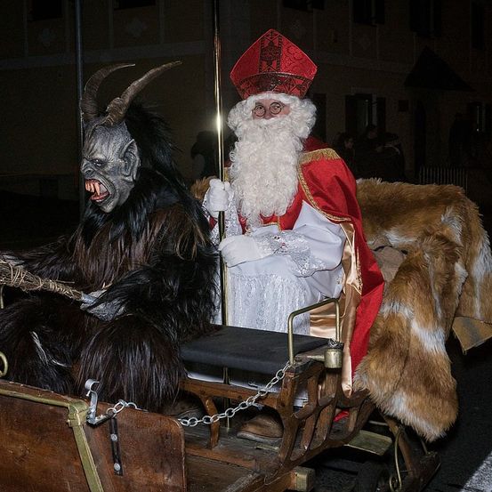 Eine Person in einem Weihnachtsmann-Kostüm fährt mit einem Mann in einem Krampus-Kostüm einen Schlitten. Sie sind draußen bei Nacht, und es gibt Menschen im Hintergrund.
