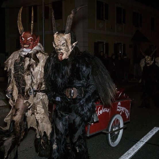 Zwei Personen in aufwendigen Kostümen mit Hörnern, Masken und dunklen Outfits gehen bei Nacht vor einem Gebäude, mit einem roten Dreirad und Zuschauern.