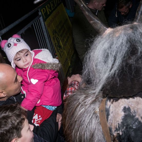 Ein Mann hält ein junges Mädchen in einem rosa Mantel, eine maskierte Person im Hintergrund. Die Umgebung scheint ein Karneval oder Festival zu sein.