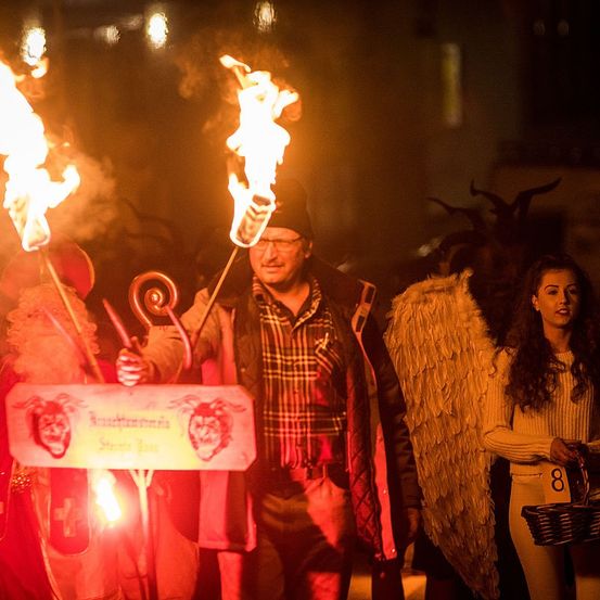 Bei Nacht hält ein Mann brennende Fackeln. Hinter ihm steht eine Frau mit Engelsflügeln und einem Korb. Sie sind von anderen in Kostümen umgeben, vielleicht für eine Halloween-Feier.