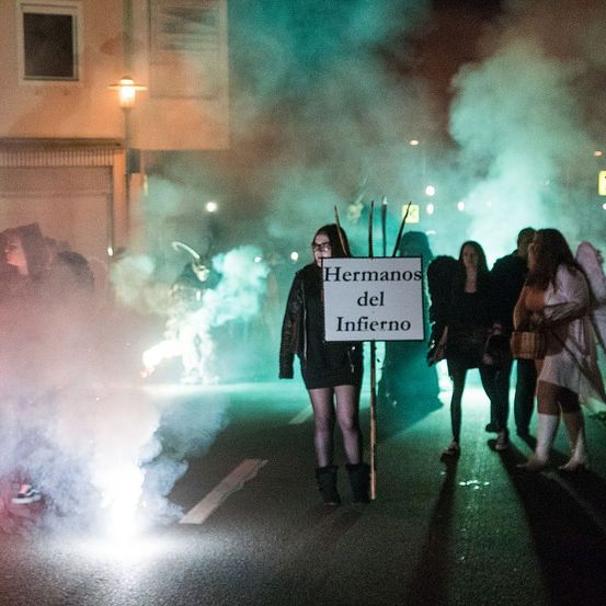 Eine Frau, die ein Schild mit der Aufschrift Hermanos del Infierno hält, steht inmitten von Rauch und anderen Demonstranten, möglicherweise während eines Halloween-Events.