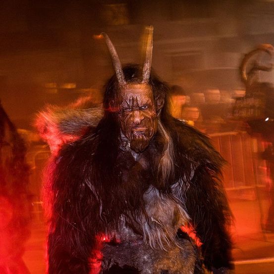 Eine Person mit einer gehörnten Maske steht unter rotem Licht, wahrscheinlich bei einer Halloween-Veranstaltung. Die Maske bedeckt das Gesicht, und sie trägt ein pelziges Kostüm. Verschwommene Figuren im Hintergrund deuten auf eine volle Umgebung hin.