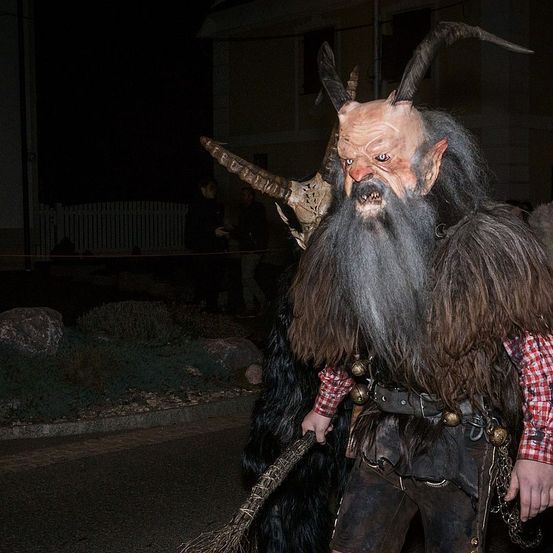 Eine Person in einer gehörnten Maske und Fellkostüm steht nachts auf der Straße, eine Besen in der Hand. Dahinter gehen zwei Leute. Links steht ein weißer Zaun.