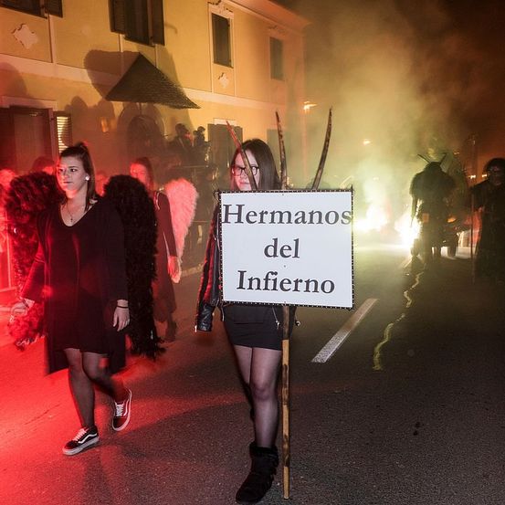 Zwei Frauen in Kostümen gehen eine Straße entlang mit einem Schild, das 'Hermanos del Infierno' steht, andere Leute in Kostümen in der Nähe. Rauch und Lichter im Hintergrund.